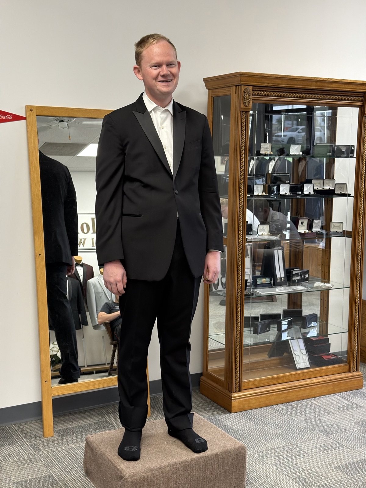 Nick Thompson grinning in a black tuxedo at his fitting, standing on the tailor's pedestal in his socks.